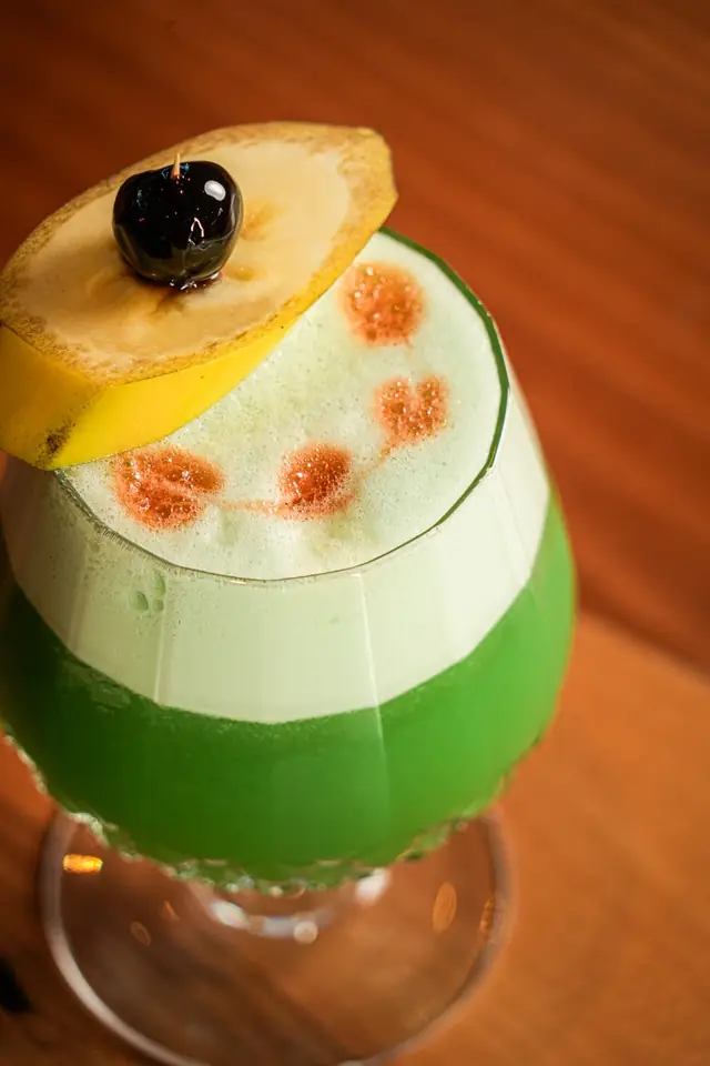 A bright green cocktail topped with foam, a lemon slice and a cherry